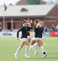 Three women in black athletic shirts and white shorts are on a grassy field, engaged in some activity that appears to be related to sports practice or warm-up. They are in an open area with a building with large windows in the background. There are also a few people visible near the building. The field is marked for a sporting event, and a few pieces of sports equipment can be seen, including a soccer ball and some training cones.