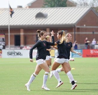 Three women in black athletic shirts and white shorts are on a grassy field, engaged in some activity that appears to be related to sports practice or warm-up. They are in an open area with a building with large windows in the background. There are also a few people visible near the building. The field is marked for a sporting event, and a few pieces of sports equipment can be seen, including a soccer ball and some training cones.