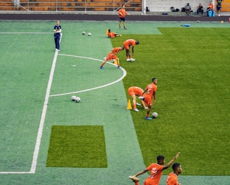 A group of people wearing bright orange sportswear are practicing on a green field. They appear to be athletes, likely soccer players, stretching and preparing for training. There are several soccer balls on the field, and a coach is observing from a distance. The field is surrounded by bleachers with a few onlookers seated, and the area is bordered by trees and residential buildings in the background.