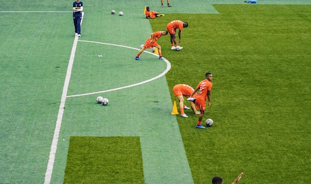 A group of people wearing bright orange sportswear are practicing on a green field. They appear to be athletes, likely soccer players, stretching and preparing for training. There are several soccer balls on the field, and a coach is observing from a distance. The field is surrounded by bleachers with a few onlookers seated, and the area is bordered by trees and residential buildings in the background.