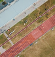 An aerial view of a sports complex featuring a running track and a grassy field. The tracks are red and situated next to a large building. There are several people scattered across the area, some on the track and others near the building. The surroundings include trees and geometric pathways, adding structure to the scene.