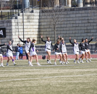 A group of athletes warming up or practicing on a grassy field near a track. They are wearing athletic gear, including shirts and shorts, and appear to be involved in a group exercise or routine. Most have one arm raised, and they are positioned in a line with a large stone wall in the background. A sign on the wall reads 'CAA', indicating a sports conference or event.