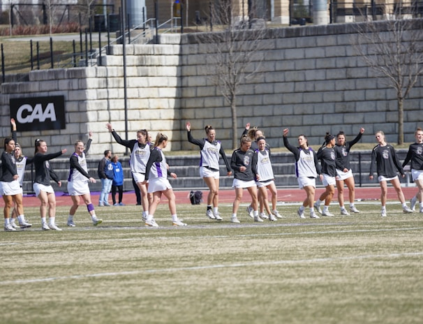 A group of athletes warming up or practicing on a grassy field near a track. They are wearing athletic gear, including shirts and shorts, and appear to be involved in a group exercise or routine. Most have one arm raised, and they are positioned in a line with a large stone wall in the background. A sign on the wall reads 'CAA', indicating a sports conference or event.