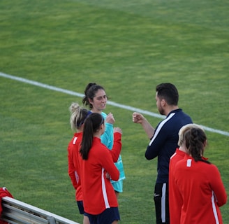 A group of people wearing sports attire are gathered on a grassy field. Some individuals are dressed in red jackets, while others are in turquoise and navy blue. A coach appears to be giving instructions. Sports equipment, such as water bottles and a bench with towels, can be seen nearby.