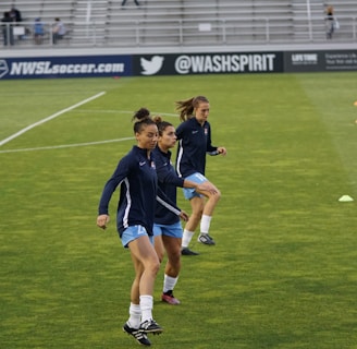 Three athletes are warming up on a grassy field, each wearing navy blue jackets and light blue shorts. They are engaged in a drill involving small orange and blue cones placed on the field. Spectators can be seen sitting in the background in a stadium setting, and advertisements are displayed on the side of the field.