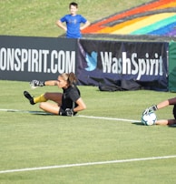 Two football players are performing a training drill on a grassy field. One is leaning back while controlling a soccer ball with her hands, and the other is lying on her back with her legs and arms up, holding another soccer ball. Both are wearing black attire with yellow socks. In the background, banners display social media handles and web addresses, and a person in a blue shirt stands nearby, watching the scene.