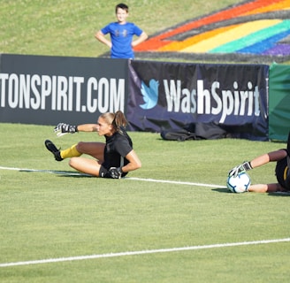 Two football players are performing a training drill on a grassy field. One is leaning back while controlling a soccer ball with her hands, and the other is lying on her back with her legs and arms up, holding another soccer ball. Both are wearing black attire with yellow socks. In the background, banners display social media handles and web addresses, and a person in a blue shirt stands nearby, watching the scene.