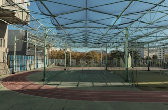 A spacious indoor sports facility with a tennis court and running tracks. The court is surrounded by high nets and tall, green metal structures supporting a roof. The area is well-lit with natural light filtering in through a transparent ceiling. In the background, there are trees with autumn foliage and several residential buildings.