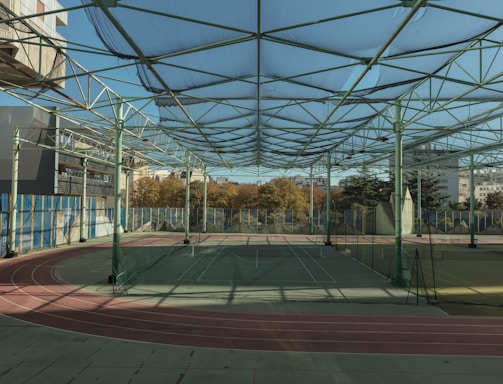 A spacious indoor sports facility with a tennis court and running tracks. The court is surrounded by high nets and tall, green metal structures supporting a roof. The area is well-lit with natural light filtering in through a transparent ceiling. In the background, there are trees with autumn foliage and several residential buildings.