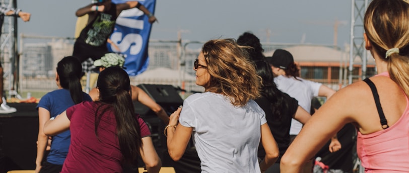 A group of people appears to be participating in an outdoor fitness class or dance session. They are dressed in athletic clothing, and a blue banner can be seen in the background, along with a stage. The setting suggests a lively and energetic atmosphere.