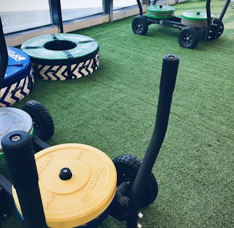 A gym setting with equipment used for weight training resting on artificial grass. There are weighted sleds with handles, featuring colorful plates stacked on them, including yellow and green ones. Additional training apparatuses like large round cushions or bumpers with blue and green covers are visible, designed for strength workouts. The room is bordered by large windows allowing natural light to enter.