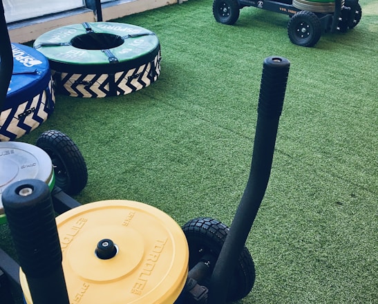 A gym setting with equipment used for weight training resting on artificial grass. There are weighted sleds with handles, featuring colorful plates stacked on them, including yellow and green ones. Additional training apparatuses like large round cushions or bumpers with blue and green covers are visible, designed for strength workouts. The room is bordered by large windows allowing natural light to enter.