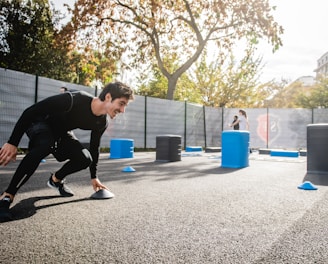 A man in athletic clothing is engaged in an outdoor workout or training session, bent forward while holding a marker or cone. The environment includes other sports equipment such as blue and gray barrels and more cones scattered around. In the background, additional people are seen participating, with trees and a building visible, suggesting a park or recreational area.