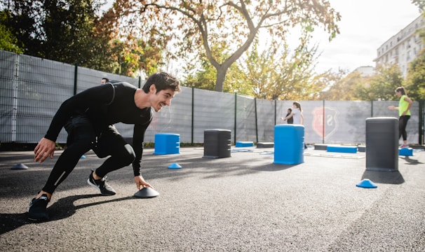 A man in athletic clothing is engaged in an outdoor workout or training session, bent forward while holding a marker or cone. The environment includes other sports equipment such as blue and gray barrels and more cones scattered around. In the background, additional people are seen participating, with trees and a building visible, suggesting a park or recreational area.