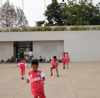 Several children in red sports uniforms are actively participating in a cone drill on a large concrete surface. The background features a modern white building, trees, and a few people sitting or standing near the structure.