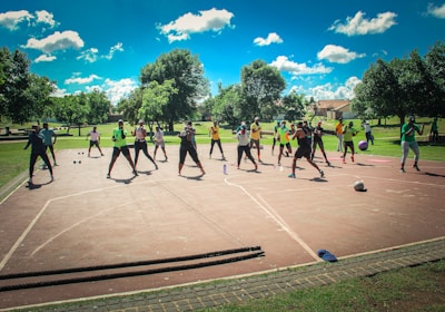 A group of people are exercising outdoors on a concrete court, surrounded by green spaces and trees. They are standing in a formation, appearing to engage in an organized fitness class. Each participant wears athletic clothing, and several carry exercise equipment such as dumbbells and ropes. The sky is bright blue with scattered fluffy clouds, creating a vibrant and energetic atmosphere.