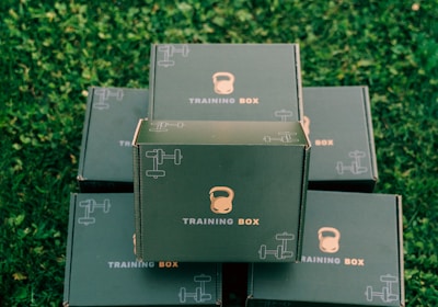 Several black boxes labeled 'Training Box' are stacked on a grassy surface. The top box features illustrations of a kettlebell and a dumbbell.