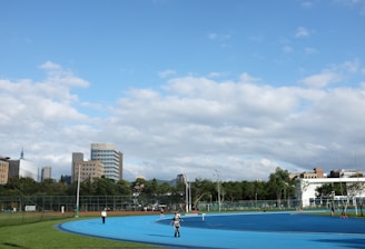 A bright outdoor sports complex features a blue running track, surrounded by green grass and trees. Several people are visible, engaged in activities on the track. In the background, urban high-rise buildings rise against a partly cloudy sky.