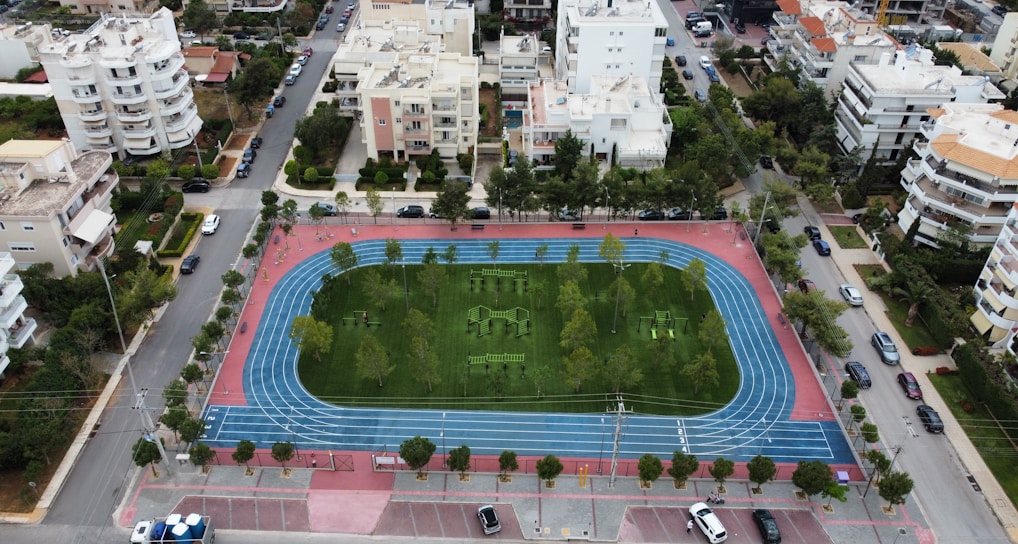 An aerial view of a sports complex featuring a blue running track surrounding a green field with workout equipment and trees. The area is bordered by residential buildings and streets with parked cars and greenery.