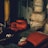 A dimly lit gym or training room featuring a pair of red boxing gloves resting on a surface. The background includes a wrapped pillar and a boxing bag, suggesting an atmosphere of sports training.